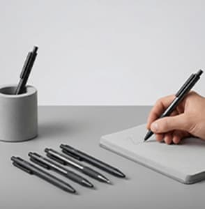 A hand writing in a notebook with various pens on the table.